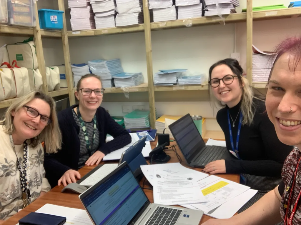 Four people sitting around a table with laptops and paperwork smiling at the camera.