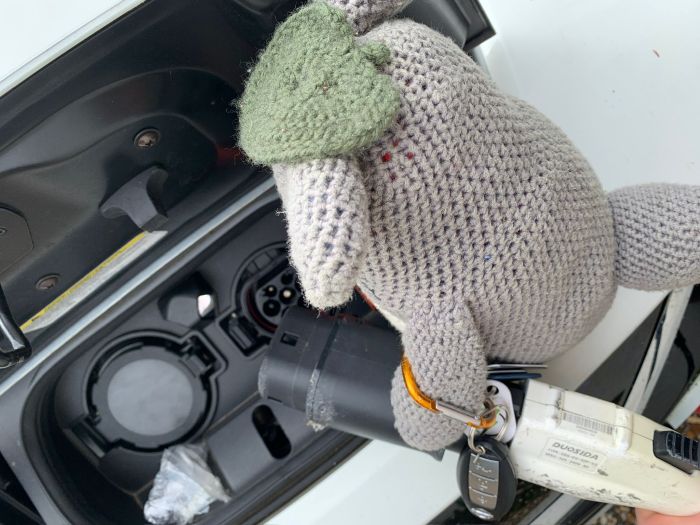 Crocheted Totoro holds a set of keys and an EV charging connector near an electric vehicle charging port.