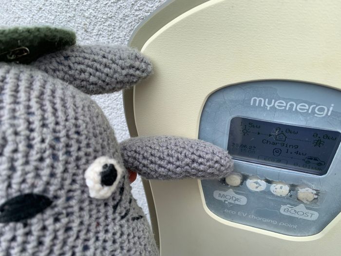 Crocheted Totoro interacts with an electric vehicle charging station displaying "Charging" at 1.4 kW.