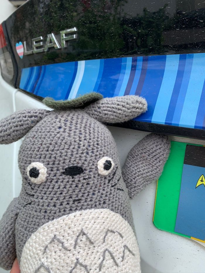 Crocheted Totoro toy poses next to the back of a white Nissan Leaf, featuring a stripes of blue (part of the warming stripes) and a pride sticker.