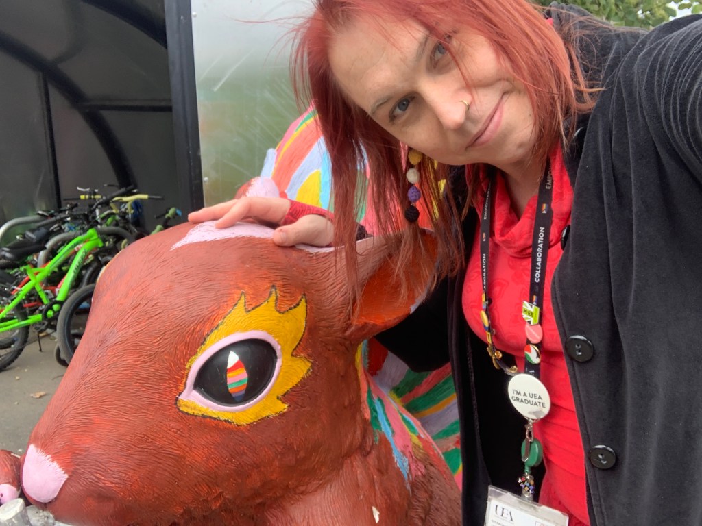 Selfie of me wearing my UEA work lanyard with my hand on the top of a giant squirrel statue with rainbow eyes and yellow painted flair around it.